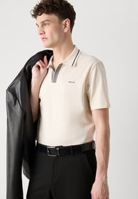 Beige Poloshirt mit gestreiftem Kragen, GANT-Logo. Modell trägt eine schwarze Lederjacke über der Schulter, kombiniert mit schwarzen Hosen und einem Gürtel.
