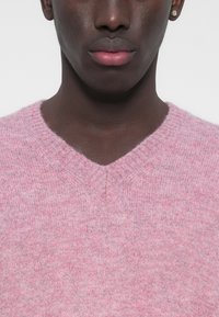 Világos rózsaszín V-nyakú pulóver puha, texturált kötött anyagból, finom melírozott mintával és bordázott gallérral.