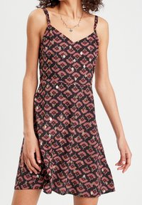 Femme portant une robe noire sans manches avec des motifs floraux roses et verts, et un collier avec un pendentif doré, debout devant un fond blanc.