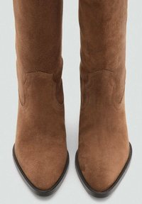 Botas de cowboy de gamuza marrón con punta afilada, que presentan costuras decorativas y una suela negra lisa. La parte superior tiene un diseño estructurado.