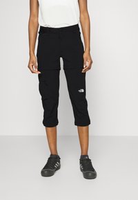 Person klädd i svarta North Face byxor som kan konverteras till capri, tillsammans med svarta Adidas sneakers, står mot en enkel vit bakgrund.