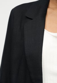 Κοντινό πλάνο ενός ατόμου που φοράει μαύρο σακάκι πάνω από λευκό ριμπ πλεκτό τοπ, δείχνοντας την περιοχή του γιακά και του ώμου.