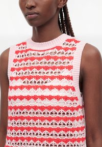 Femme portant un haut crocheté sans manches rayé rose et rouge, présentant des motifs ajourés et texturés détaillés.