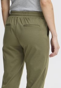 Olijfgroene stoffen broek met elastische tailleband, voorzien van twee achterzakken en een gladde textuur. Zijnaden zichtbaar.