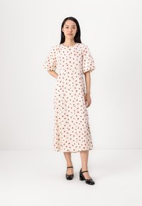 Crème midi-jurk met een rood bloemmotief, pofmouwen en een getailleerde taille. Model draagt zwarte hakken en staat op een neutrale achtergrond.