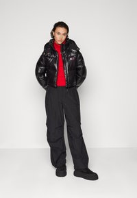 Abrigo de plumas negro con un acabado brillante, que cuenta con capucha y bolsillos delanteros, combinado con pantalones negros holgados y un suéter de cuello alto rojo. Zapatos negros.