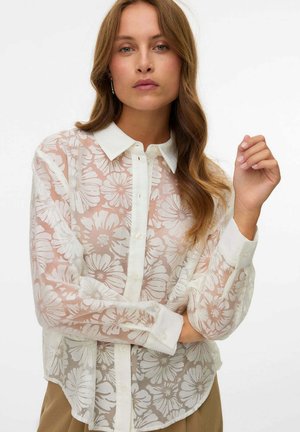 Vrouw met lang golvend haar, gekleed in een doorschijnende witte blouse met knopen en grote bloemenpatronen, gecombineerd met beige broek, tegen een effen achtergrond.
