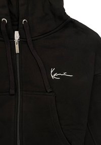 Felpa nera con zip realizzata in tessuto morbido. Presenta una tasca a canguro, cordini e un logo ricamato bianco sul petto.