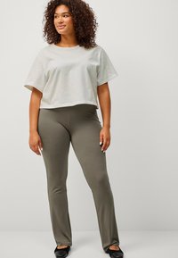 Kort, lichtgrijs T-shirt met korte mouwen gecombineerd met olijfgroene, strakke leggings. Model draagt zwarte platte schoenen en toont een casual outfit.