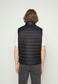 Gilet imbottito nero con colletto alto e cuciture orizzontali, realizzato in tessuto leggero. Indossato sopra una camicia bianca, che mette in risalto un look informale.