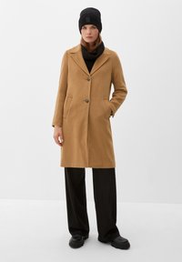 Manteau couleur camel au design cintré, avec deux poches frontales et deux boutons. Porté sur un col roulé noir et un pantalon, agrémenté d'un bonnet noir.