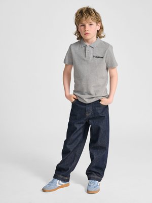 Chłopiec z rozczochranymi blond włosami stoi z rękami w kieszeniach, ubrany w szarą koszulkę polo, ciemne dżinsy i jasnoniebieskie trampki.