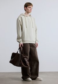 Φωτεινό μπεζ φούτερ με κουκούλα και ελαστική υφή, συνδυασμένο με χαλαρό καφέ παντελόνι. Το μοντέλο κρατάει μια μεγάλη καφέ δερμάτινη τσάντα με διπλά χερούλια.