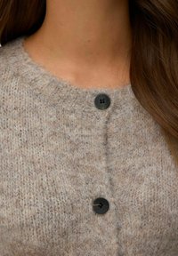 Stickad cardigan i ljusbeige med ett strukturerat mönster, försedd med två svarta knappar vid halslinningen och en rund krage.