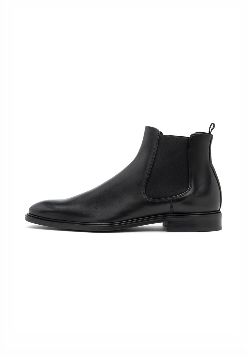 Bota de piel negra hasta el tobillo con paneles elásticos en los laterales, tira de agarre, punta redondeada y tacón plano. Textura suave y diseño minimalista.