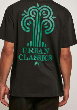 Person trägt schwarzes T-Shirt mit grünem abstraktem Baum-Design und dem Text "Urban Classics" auf der Rückseite.