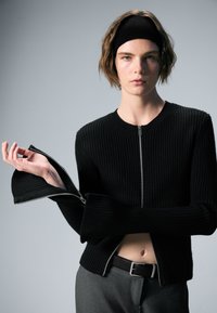 Pull en maille côtelée noire à manches longues, avec une fermeture éclair à l'avant et des poignets zippés, assorti à un pantalon gris et un bandeau noir.