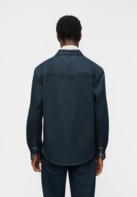 Άνδρας που φοράει σκούρο μπλε τζιν μπουφάν και τζιν παντελόνι που ταιριάζουν, φωτογραφημένος από πίσω μπροστά σε απλό ανοιχτόφωτο φόντο.