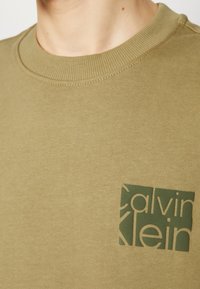 Felpa girocollo verde oliva realizzata in un tessuto morbido, con un logo testurizzato di colore verde scuro sulla parte sinistra del petto.
