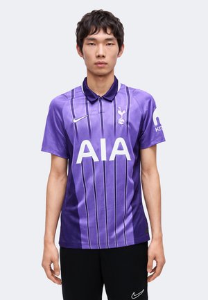 Jeune homme portant un maillot violet de Tottenham Hotspur avec le logo AIA et un short noir Nike, debout devant un fond clair unicolore.