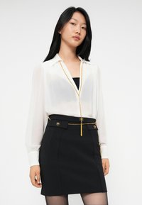 Young woman wearing sheer white blouse, black tube top, black skirt with gold chain belt and buttons, standing against white background.