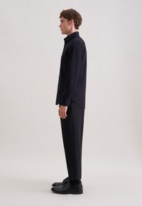 Zwart overhemd met knopen en lange mouwen, gecombineerd met op maat gemaakte zwarte broek en zwarte schoenen. Soepele stof en gestructureerd silhouet.