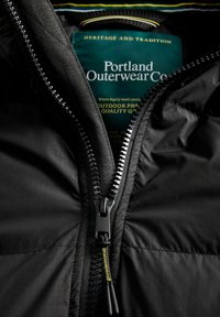 Černá náplňová bunda se zipem, texturovaná tkanina a zelený štítek s nápisem "Portland Outerwear Co." a pokyny k péči.