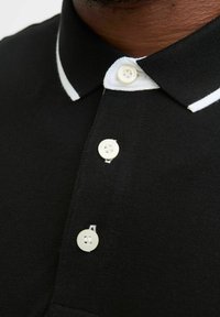 Mustakate särk valge krae ja triibuga. Omab kolme valget nööpi ja tekstuurset kangast. Lähedal vaade keskendub krae detailidele.