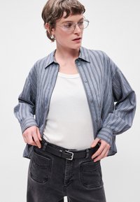 Stribet blå skjorte med lange ærmer, båret over en hvid tanktop, parret med mørkegrå jeans og et sort bælte.