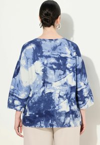 Maglione tie-dye blu e bianco con maniche a tre quarti, texture a coste e vestibilità comoda. I dettagli includono un collo rotondo e spacchi laterali.