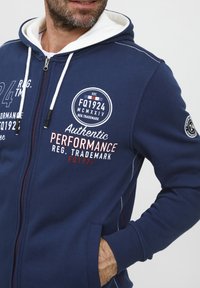 Tengerészkék cipzáras kapucnis felső, fehér béléssel, elöl piros és fehér nyomtatott szöveggel és grafikákkal, valamint oldalsó zsebekkel. Pamut keverék anyagból készült.