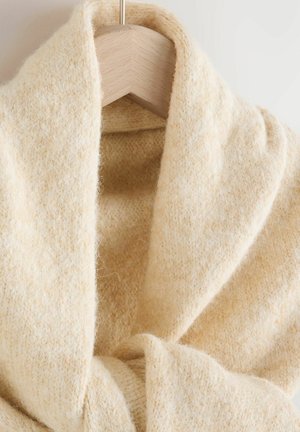 Beiger Strickschal mit einer weichen, strukturierten Oberfläche, der ein breites, fließendes Design aufweist, das elegant fällt. Hängt an einem hellen Holzkleiderbügel.