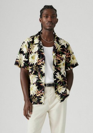 Camisa de manga corta, con cuello, en negro con un gran patrón floral en blanco y amarillo. Combinada con una camiseta sin mangas blanca y pantalones color crema.