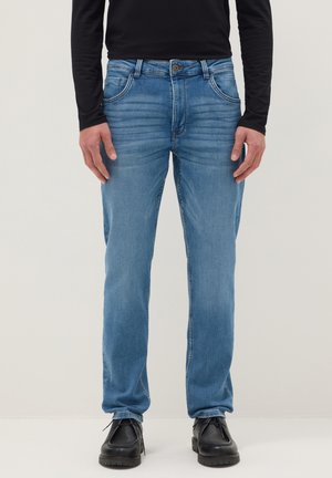 Hombre con jeans azul claro, camiseta negra de manga larga y zapatos negros con cordones, de pie frente a un fondo claro y liso.