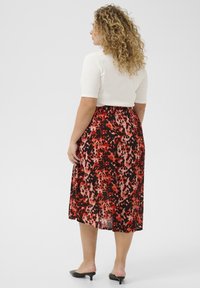 Jupe maxi en tissu fluide avec un imprimé abstrait rouge et noir, complétée par un haut ajusté blanc et des mules à talons noirs.