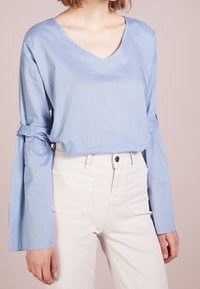 Blouse en coton bleu clair avec un décolleté en V, coupe décontractée et manches évasées avec des accents noués. Associée à un pantalon blanc taille haute.
