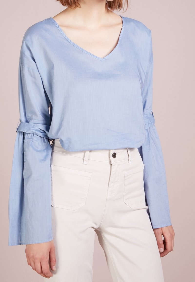 Blouse en coton bleu clair avec un décolleté en V, coupe décontractée et manches évasées avec des accents noués. Associée à un pantalon blanc taille haute.