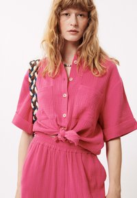Roze textiel katoenen shirt met een geknoopte taille, met korte mouwen en een knoopsluiting. Twee voorzakken zichtbaar.