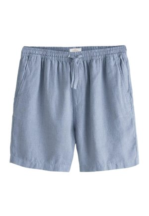 Mann trägt hellblaues Leinenhemd und Shorts, steht am Pool in einem üppigen Garten mit Bergen im Hintergrund, Hände in den Taschen, trägt Sonnenbrille.