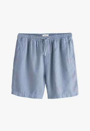 Muž v svetlomodrej ľanovej košeli a kraťasoch stojí pri bazéne v bujnej záhrade s horami v pozadí, ruky v vreckách, má na sebe slnečné okuliare.