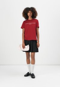 Raudona medvilninė marškinėliai su "Tommy Hilfiger" tekstu, juoda klostuota sijonas, balti iki kelių siekiantys kojinės, blizgūs juodi loaferiai, rožinė rankinė.