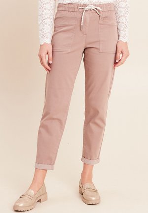 Frau trägt hellrosa umgeschlagene Hosen mit Fronttaschen, ein weißes Spitzen-Oberteil mit langen Ärmeln und beige Loafers, steht vor einem neutralen Hintergrund.