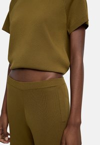 Oliwkowy, dziany crop top z krótkimi rękawami, połączony z dopasowanymi, dzianymi spodniami o wysokim stanie. Tkanina ma gładką, jednolitą teksturę.