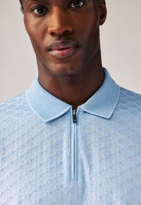 Světlemodré polo tričko s texturovaným diamantovým vzorem, krátkým zipem a měkkou, prodyšnou tkaninou. Viditelný podrobný design límce.
