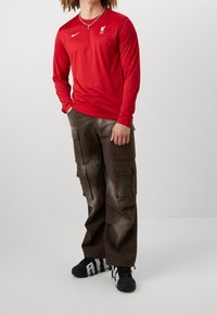 Rotes Langarmshirt mit Liverpool-Logo und Nike-Swoosh, kombiniert mit braunen Cargo-Hosen, die mehrere Taschen und eine strukturierte Oberfläche haben.