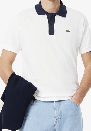 Mężczyzna ubrany w białą koszulkę polo z granatowym kołnierzem i logo Lacoste, jasnoniebieskie jeansy, trzyma granatową kurtkę w lewej ręce.
