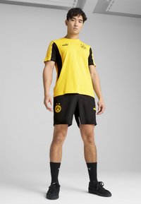 Gul og sort sportsbluse med kortærmer, logo på brystet; sorte shorts med teama-logo; sorte sokker og sko. Glat stofstruktur.
