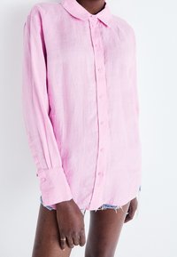 Persona con camicia rosa chiaro a maniche lunghe e bottoni, e pantaloni shorts in denim sfilacciati, in piedi di fronte a uno sfondo bianco.