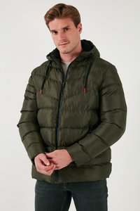 Bărbat purtând o jachetă matlasată cu glugă, de culoare verde închis, cu șnururi negre și blugi închiși la culoare, stând în picioare pe un fundal simplu și deschis la culoare.