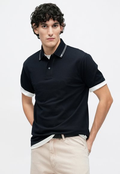 Μπλε ναυτικό πόλο μπλουζάκι με κοντά μανίκια, διακοσμημένο με λευκό γιακά και λεπτομέρειες στα μανίκια. Λείο ύφασμα, δύο κουμπιά στη λαιμόκοψη, διακριτικό λογότυπο κοντά στον γιακά.